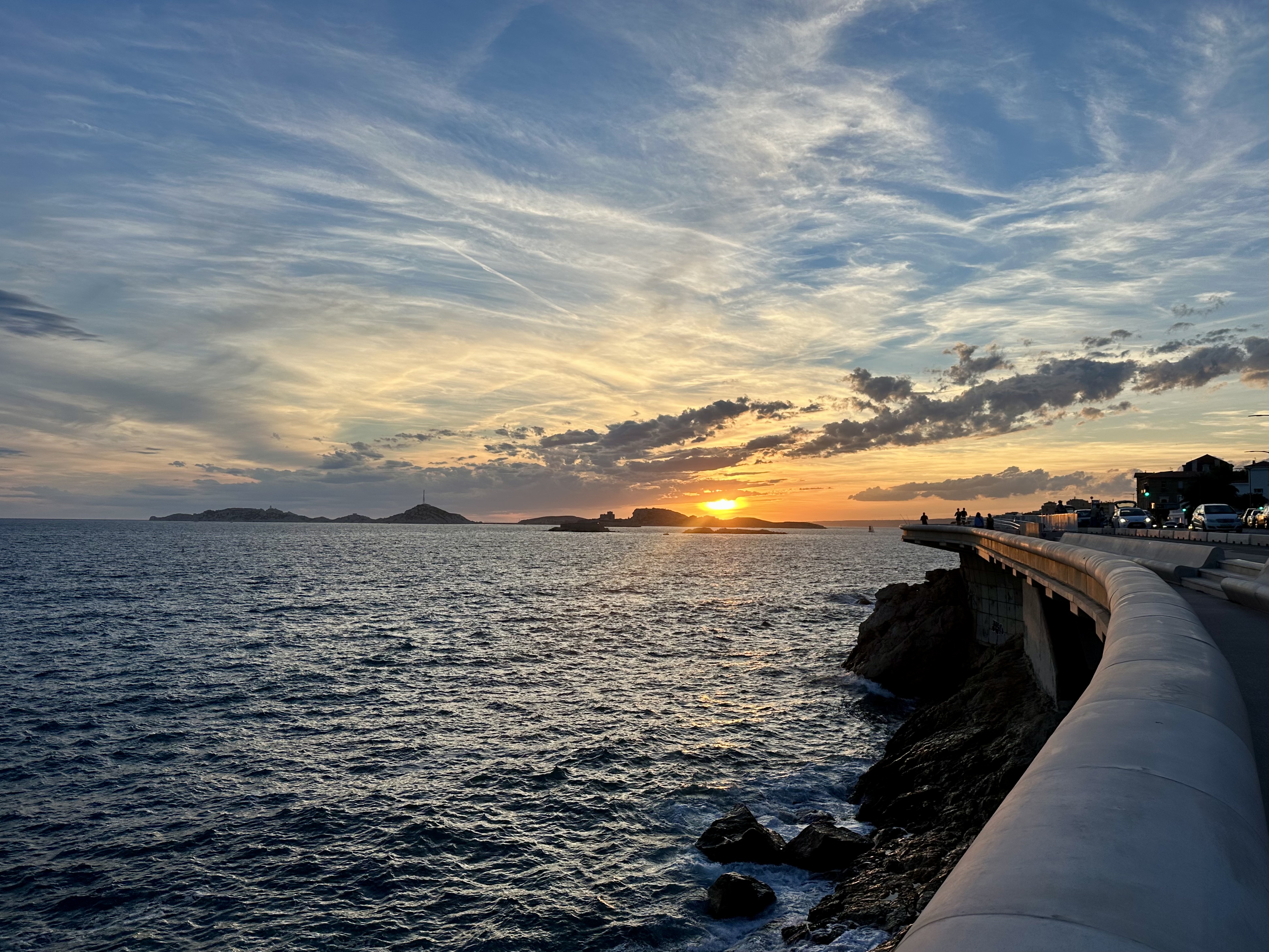 Coucher de soleil sur la mer à Marseille, avec des îles à l’horizon et une promenade longeant le rivage à droite.